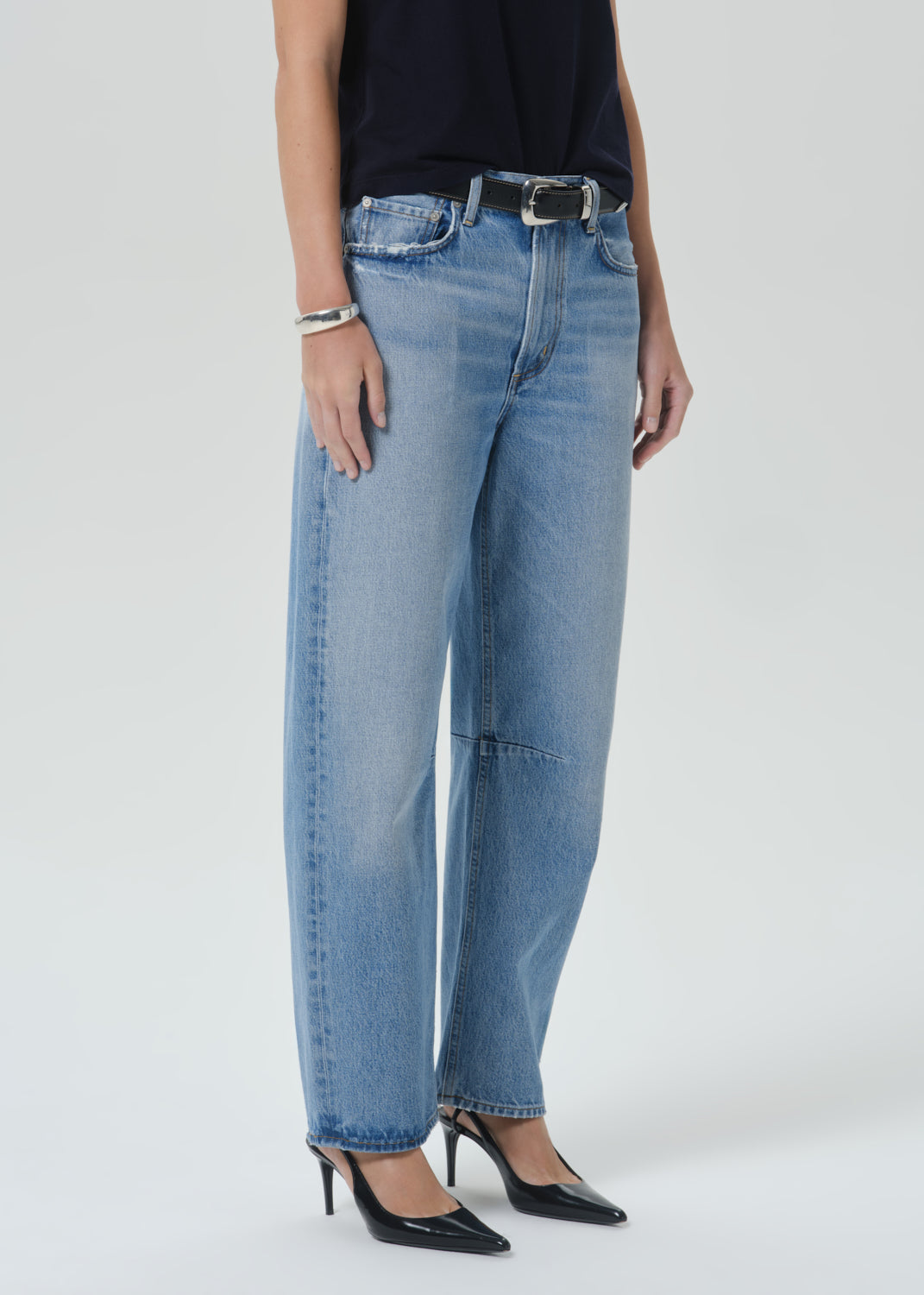 A model wearing light-wash blue wide-leg denim jeans with a black belt, paired with a black top and black pointed-toe heels, photographed against a grey background.