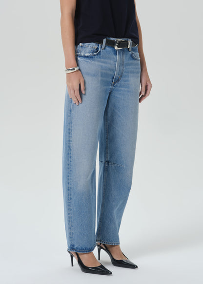A model wearing light-wash blue wide-leg denim jeans with a black belt, paired with a black top and black pointed-toe heels, photographed against a grey background.