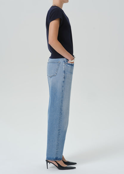 A side-profile studio shot of a model wearing light-wash blue straight-leg jeans, a dark navy short-sleeve top, and black pointed-toe heels against a grey background.