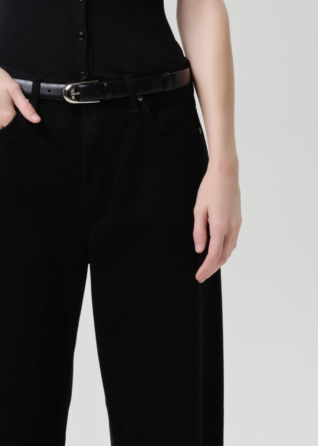 A close-up of a person wearing black wide-leg trousers and a black belt with a silver buckle, paired with a black ribbed top, against a grey background.