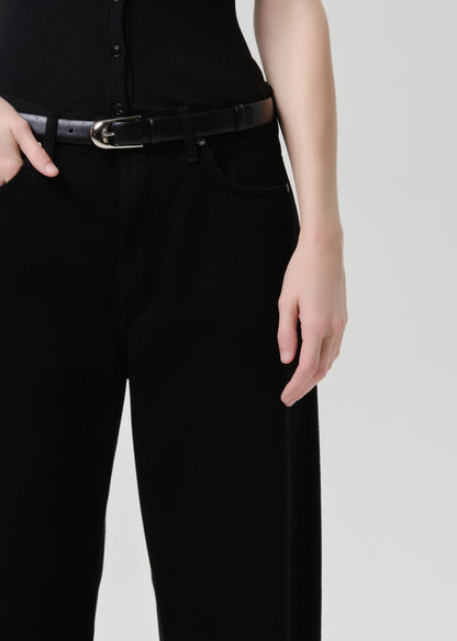 A close-up of a person wearing black wide-leg trousers and a black belt with a silver buckle, paired with a black ribbed top, against a grey background.