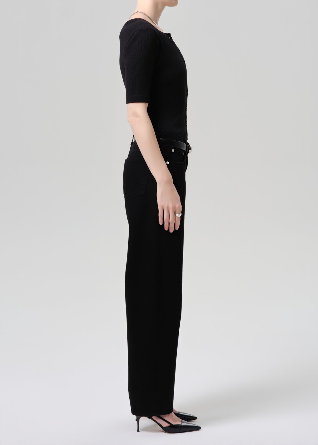 A model shown in profile wearing a black short-sleeve top and black wide-leg trousers with a black belt, paired with black pointed-toe heels, against a grey  background.