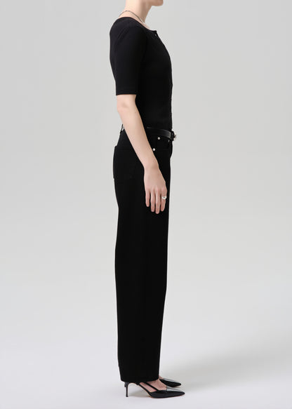 A model shown in profile wearing a black short-sleeve top and black wide-leg trousers with a black belt, paired with black pointed-toe heels, against a grey  background.