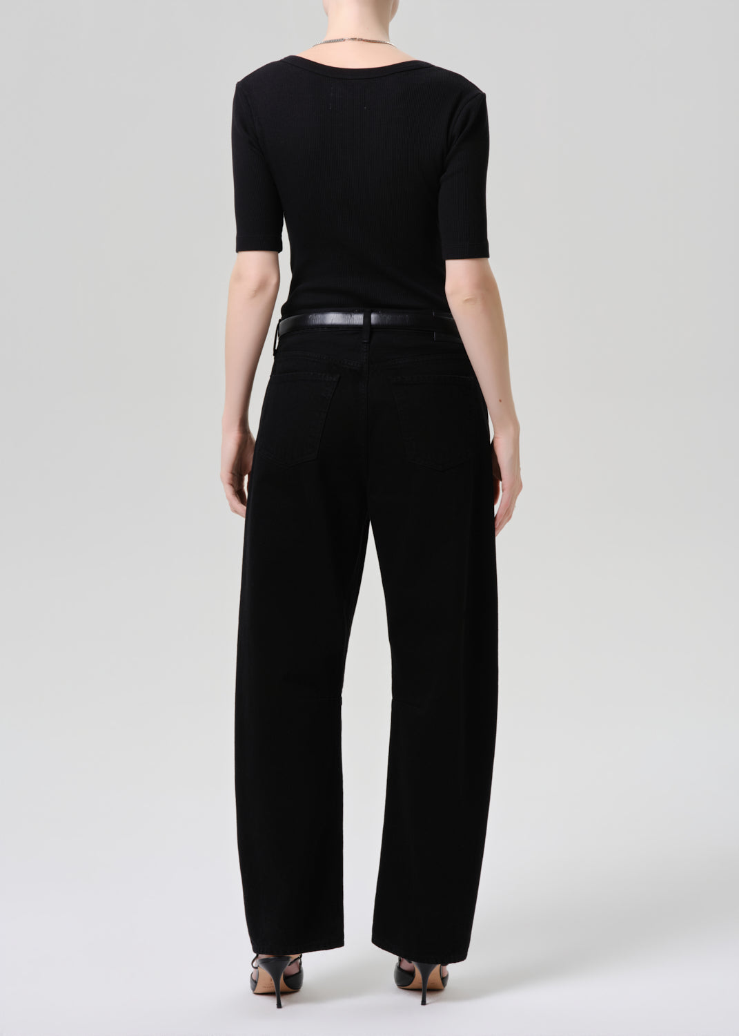 A model shown from the back wearing a black short-sleeve top, black wide-leg trousers, a thin black belt, and black pointed-toe heels against a grey background.