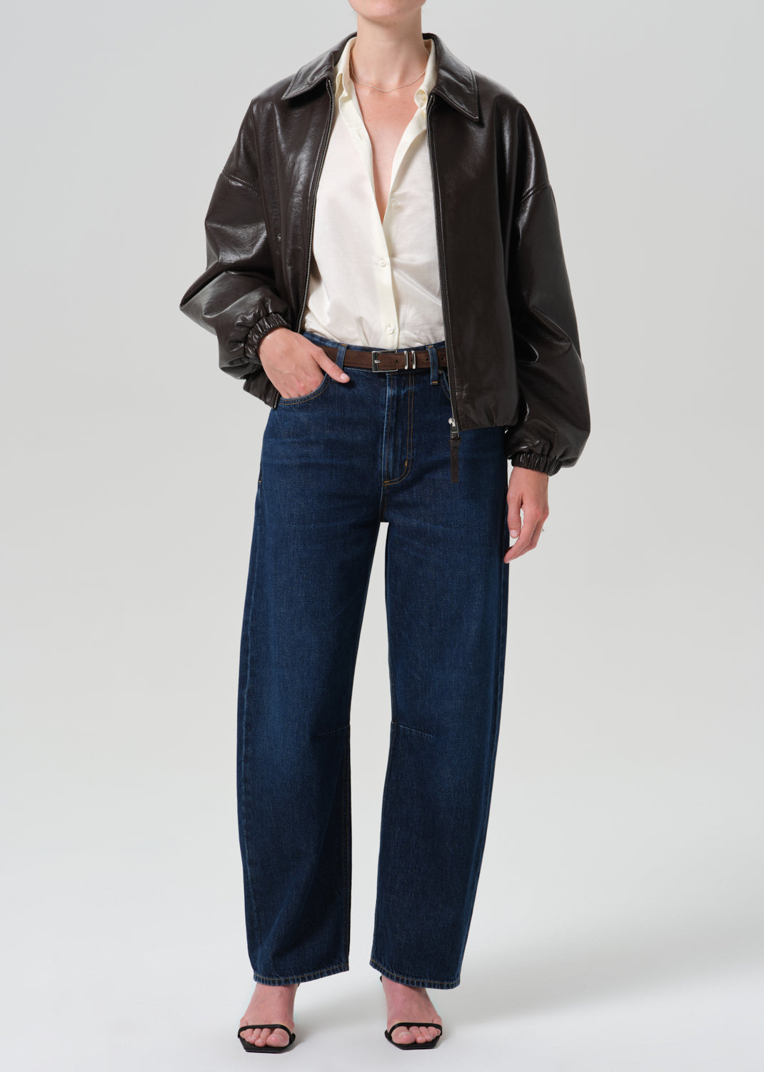 A model wearing a black faux-leather jacket, an off-white button-down shirt, dark blue wide-leg jeans, and black sandals, photographed against a grey background.