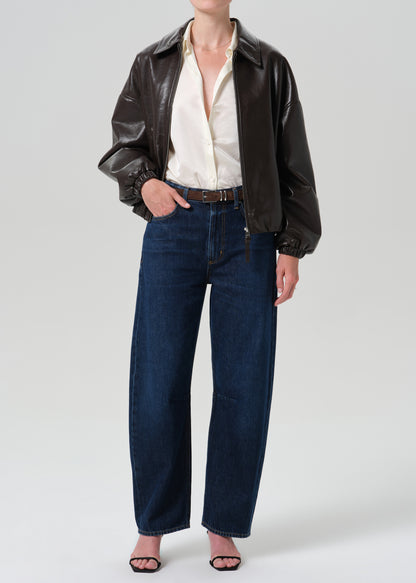 A model wearing a black faux-leather jacket, an off-white button-down shirt, dark blue wide-leg jeans, and black sandals, photographed against a grey background.