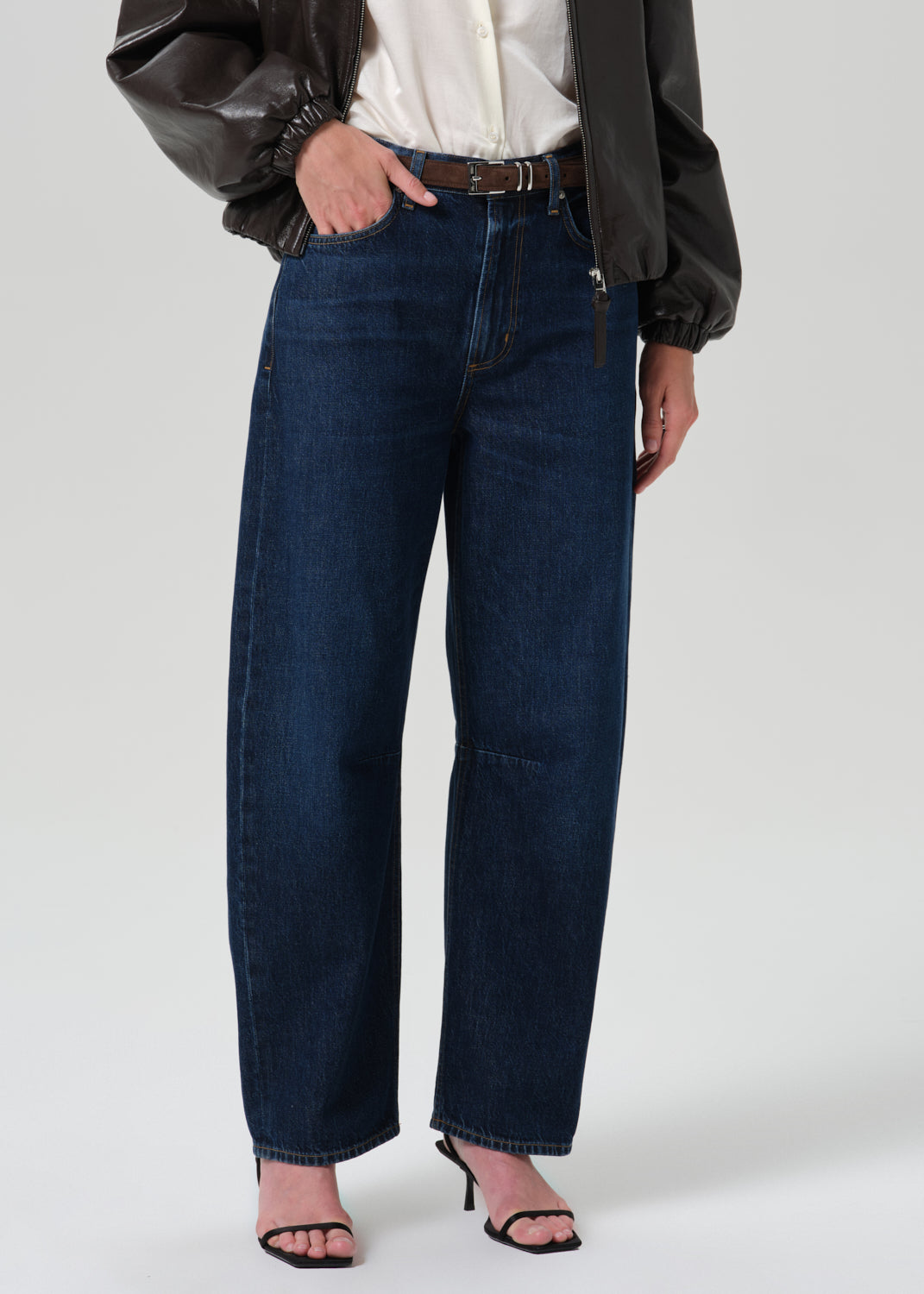 A model wearing dark blue wide-leg denim jeans, styled with a white button-up shirt, a black leather jacket, and black heeled sandals, photographed against a grey background.