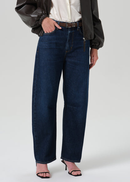 A model wearing dark blue wide-leg denim jeans, styled with a white button-up shirt, a black leather jacket, and black heeled sandals, photographed against a grey background.