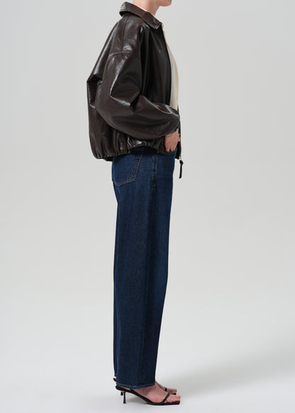 A model shown from the side wearing a dark brown faux-leather jacket, dark blue wide-leg jeans, and black heeled sandals against a grey background.