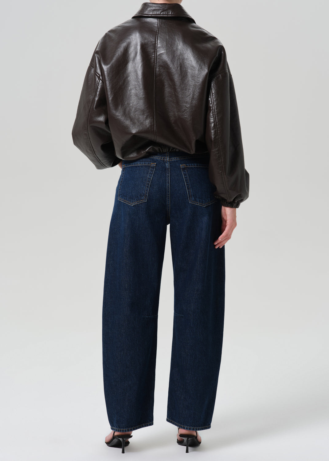 A model shown from the back wearing a dark brown leather jacket and dark blue wide-leg denim jeans, paired with black heels, against a grey  background.