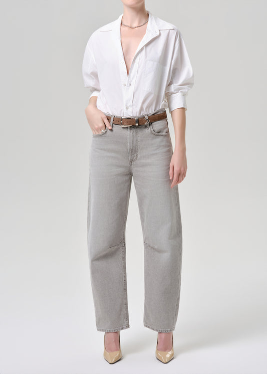 A model wearing light grey wide-leg jeans, a white button-down shirt, a brown leather belt, and beige heels, photographed against a grey background.