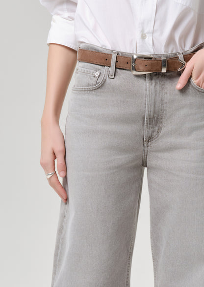 Close-up of a person wearing light grey wide-leg jeans with a brown leather belt, a white button-up shirt, and silver rings against a grey background.