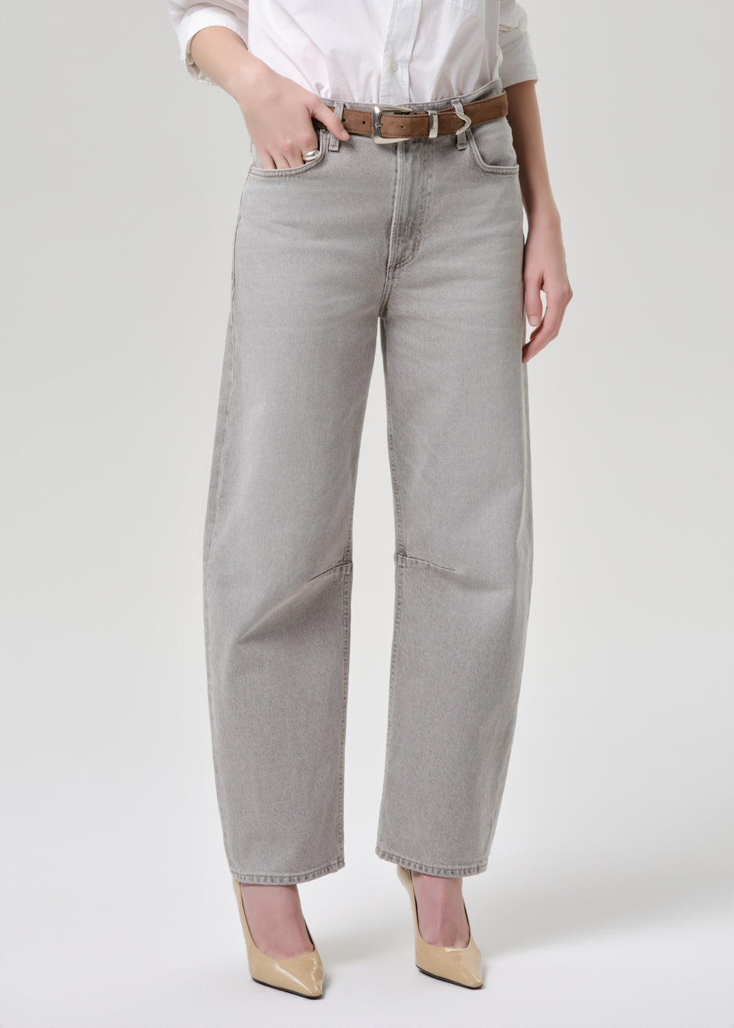A model wearing light grey wide-leg cropped jeans with a brown belt, styled with a white button-down shirt and beige pointed-toe heels against a light grey studio background.