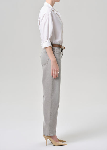 A side-profile studio shot of a model wearing light grey straight-leg jeans, a white button-down shirt, a brown belt, and beige heels against a light grey background.