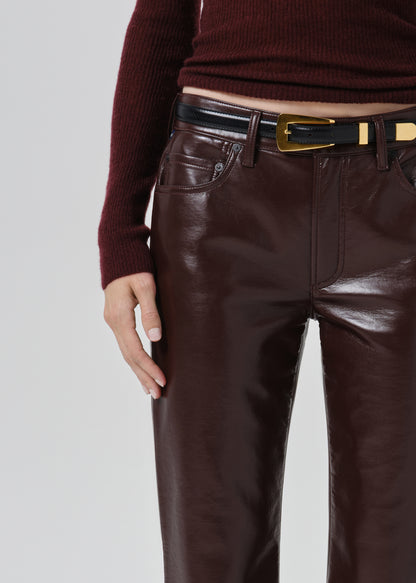 Close-up of a person wearing dark brown faux-leather trousers with a black leather belt featuring a gold buckle, paired with a burgundy ribbed knit top against a grey background.