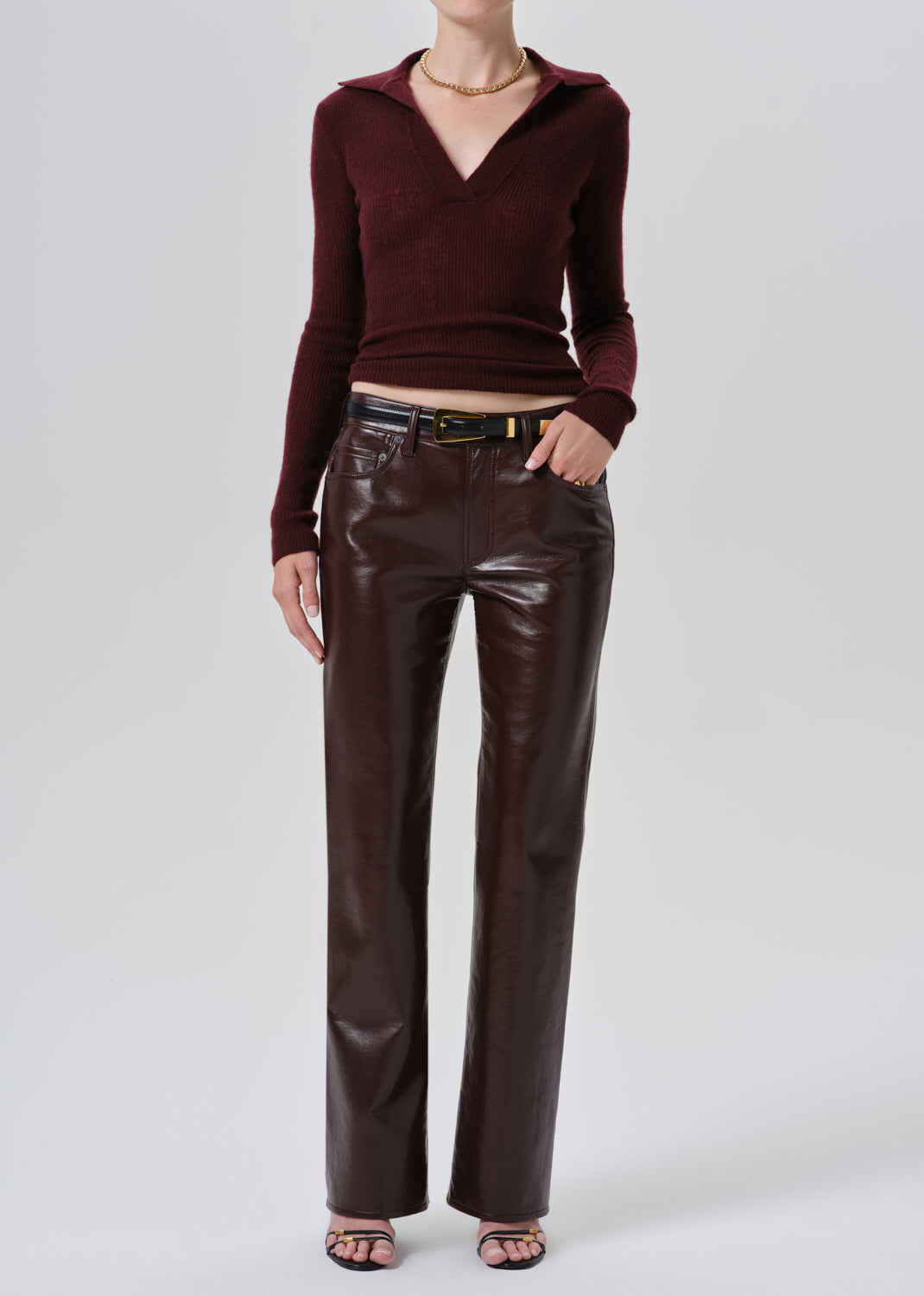 A model wearing a dark burgundy ribbed knit polo-neck long-sleeve top and matching dark brown faux-leather wide-leg trousers, styled with a black belt and metallic buckle, against a grey background.