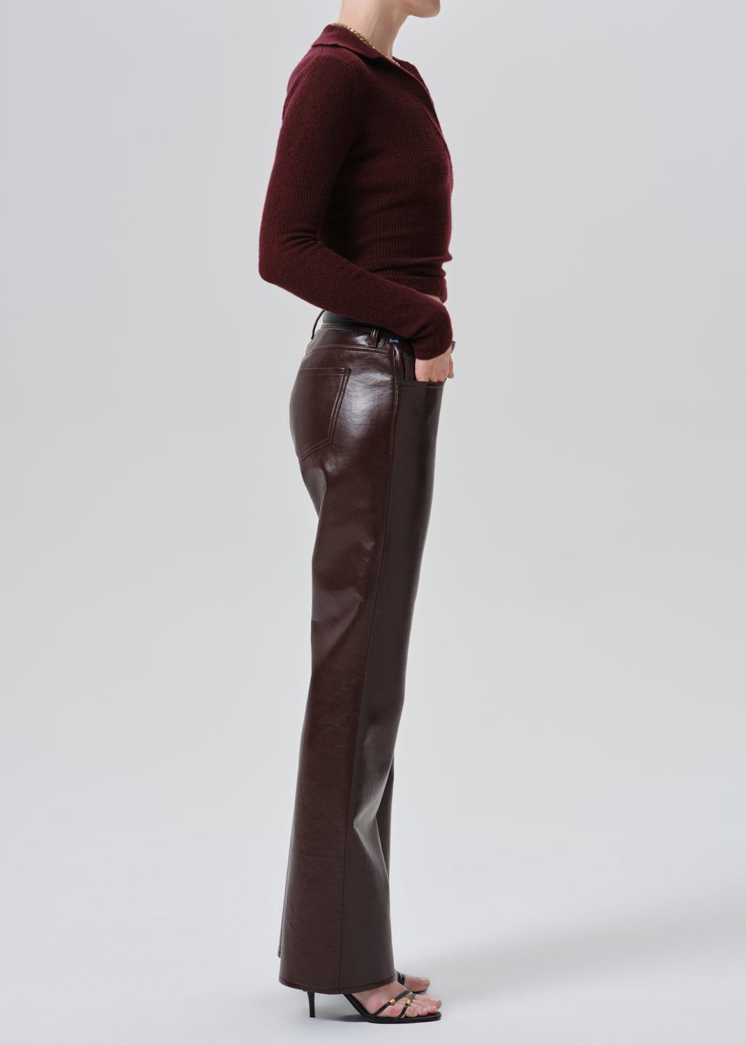 A model shown from the side wearing a dark burgundy long-sleeve mock-neck top and dark brown faux-leather flare pants, paired with black heeled sandals, against a grey background.