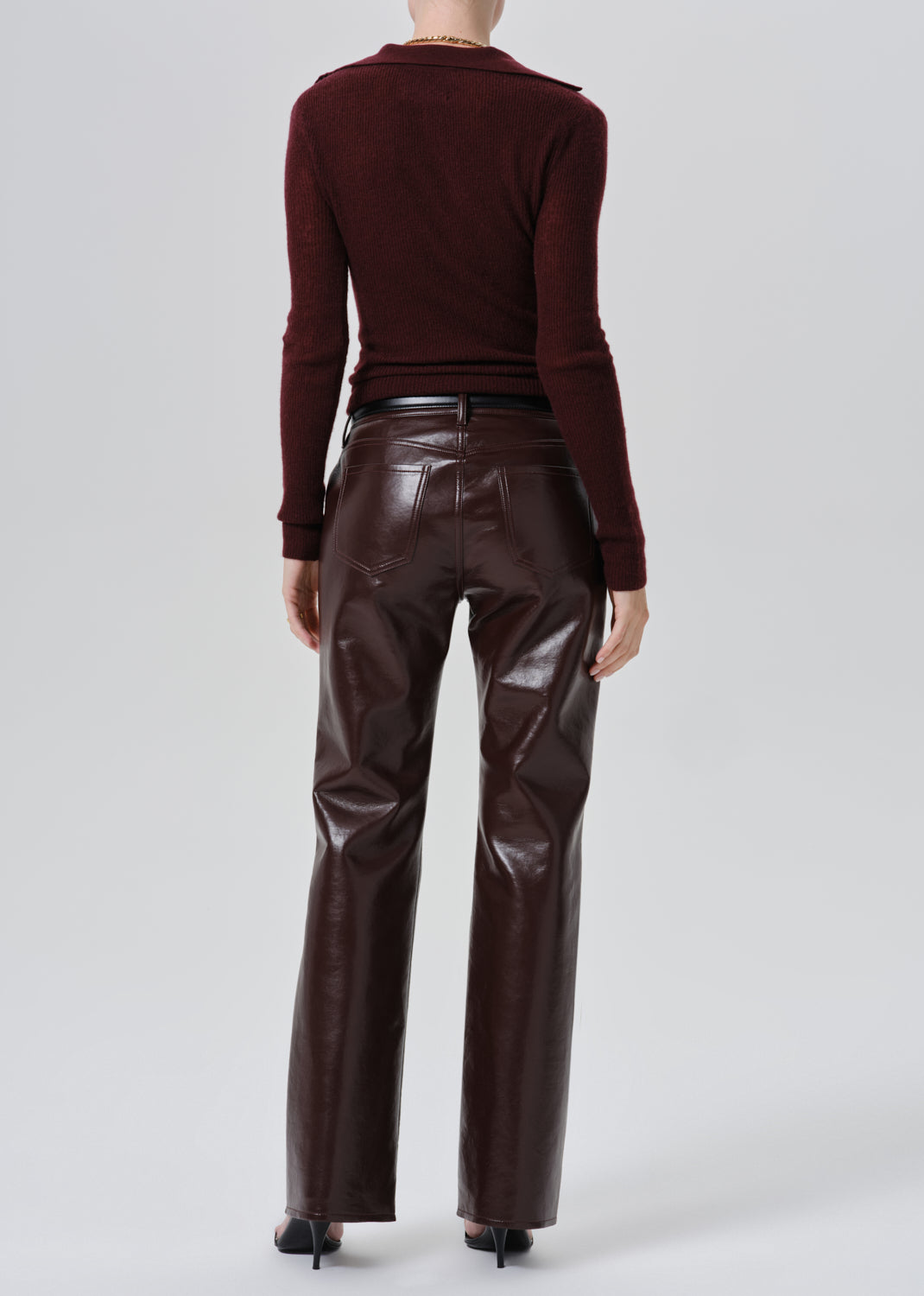 A model shown from the back wearing a dark burgundy long-sleeve top and shiny dark brown faux-leather pants against a grey background.