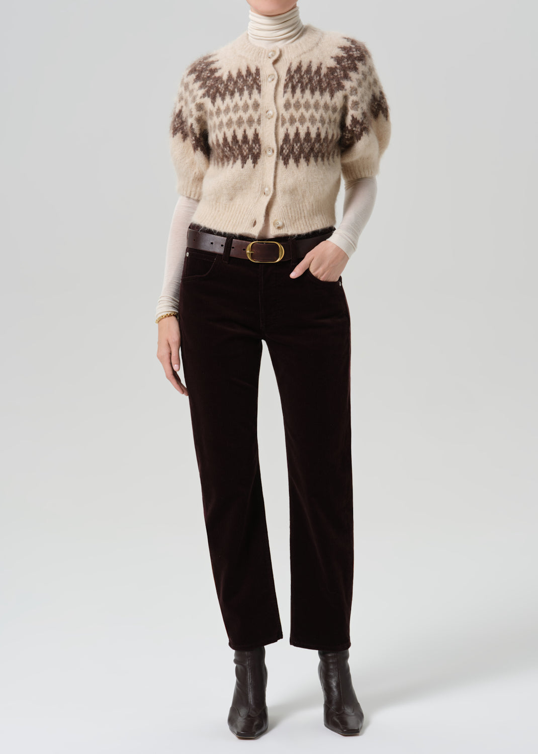 Person wearing dark brown straight leg jeans with a cream patterned short sleeve sweater on a grey background