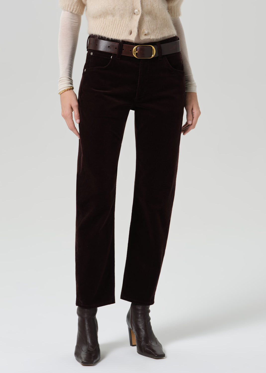Person wearing dark brown straight leg jeans with a cream patterned short sleeve sweater and black ankle boots on a grey background