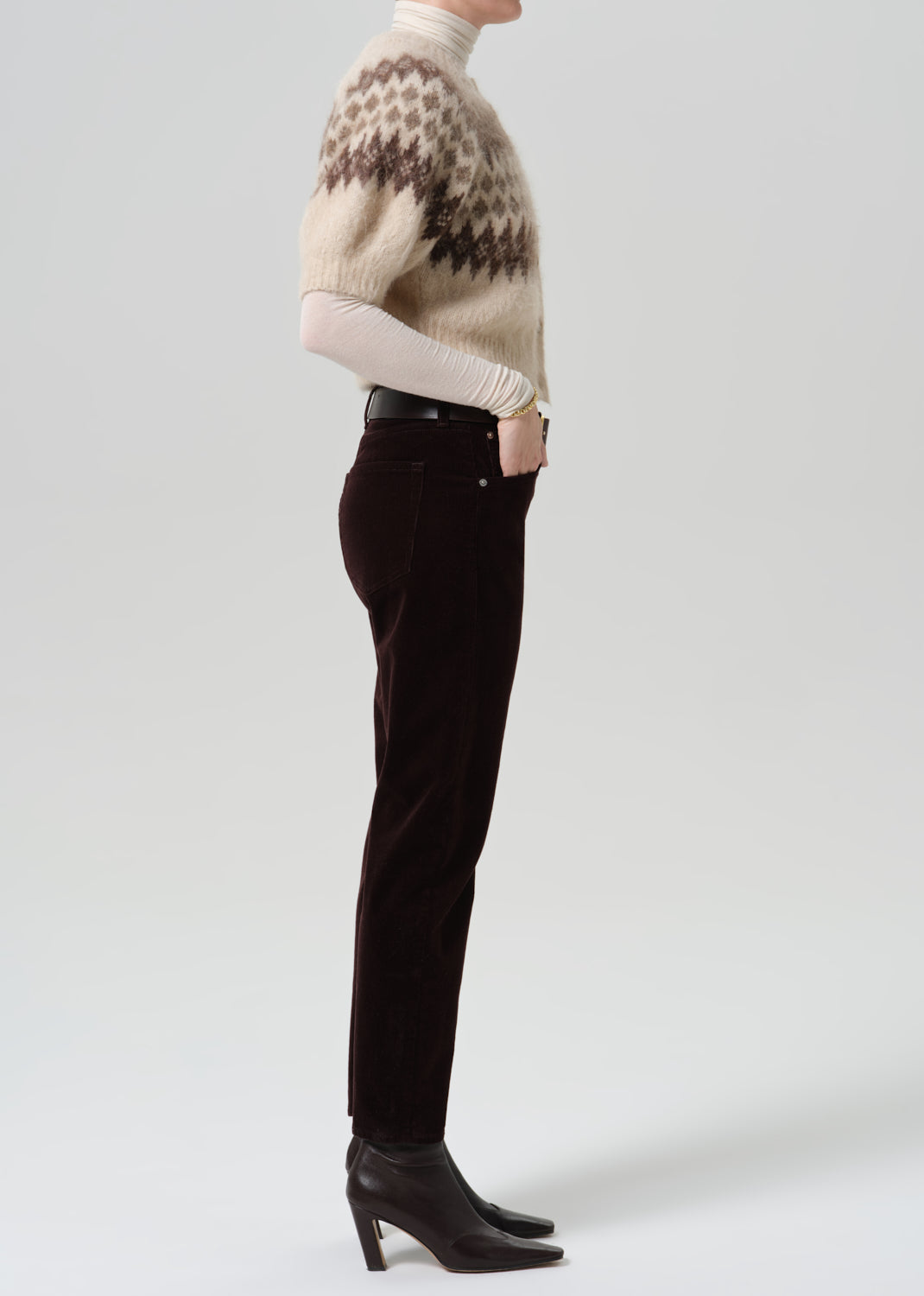 Person wearing dark brown straight leg jeans with a cream patterned short sleeve sweater viewed from the side on a grey background