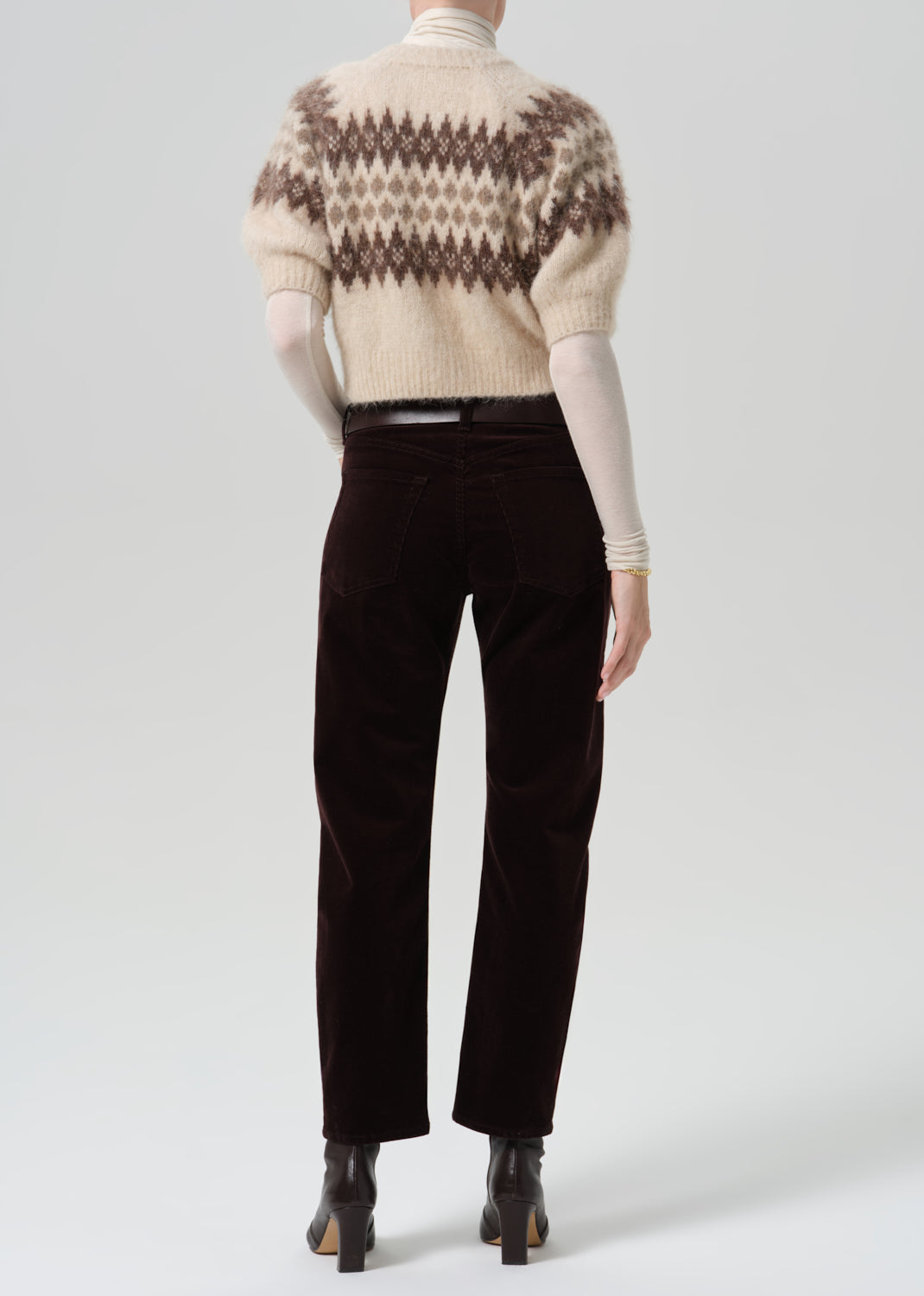 Person wearing dark brown straight leg jeans with a cream patterned short sleeve sweater viewed from the back on a grey background