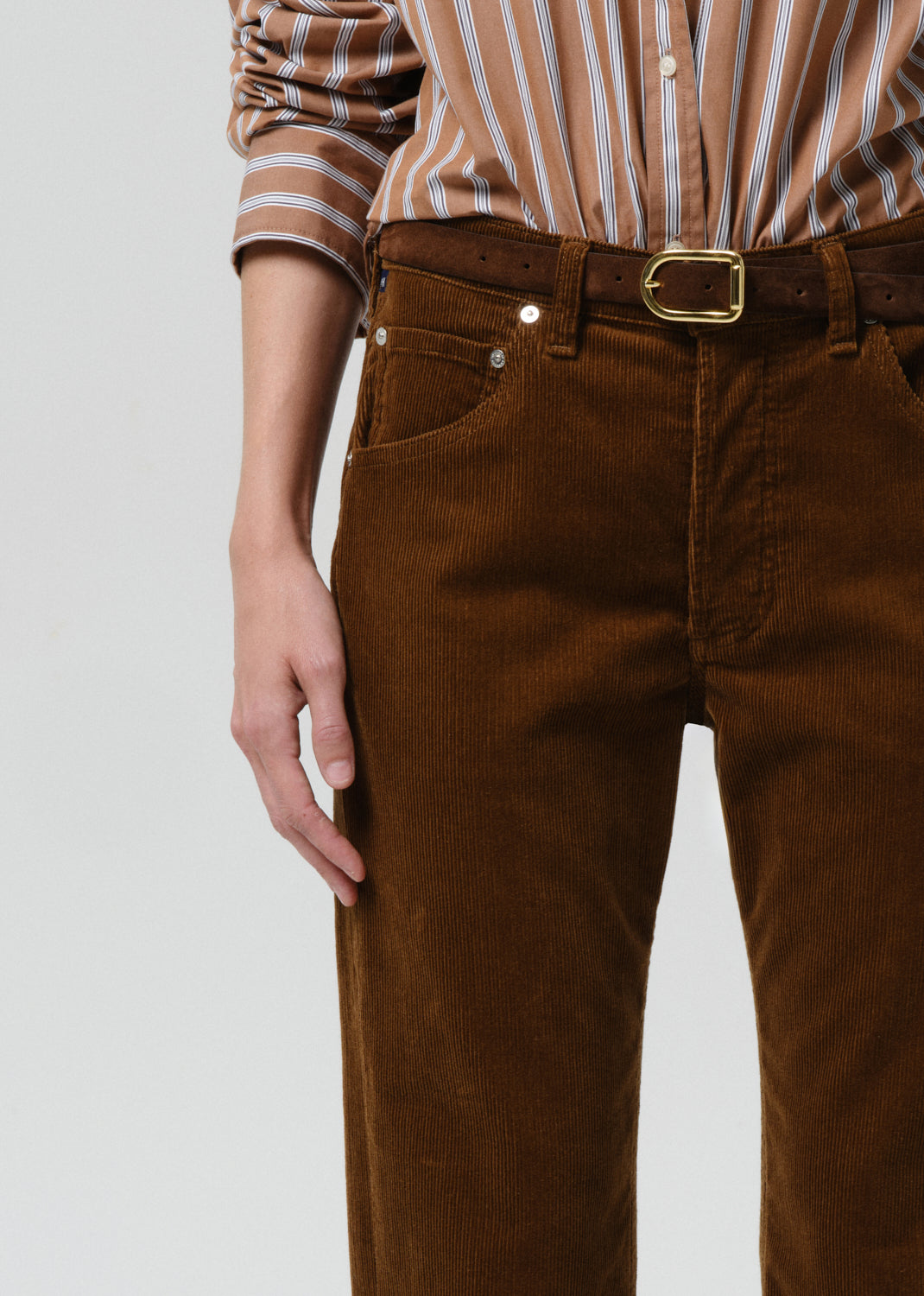 Brown corduroy pants with a gold belt buckle worn by a person on a grey background