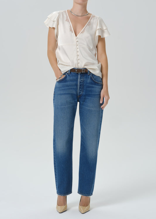 A model wearing a cream ruffled short-sleeve blouse and blue straight-leg jeans with a brown belt, paired with beige heels, against a grey background.