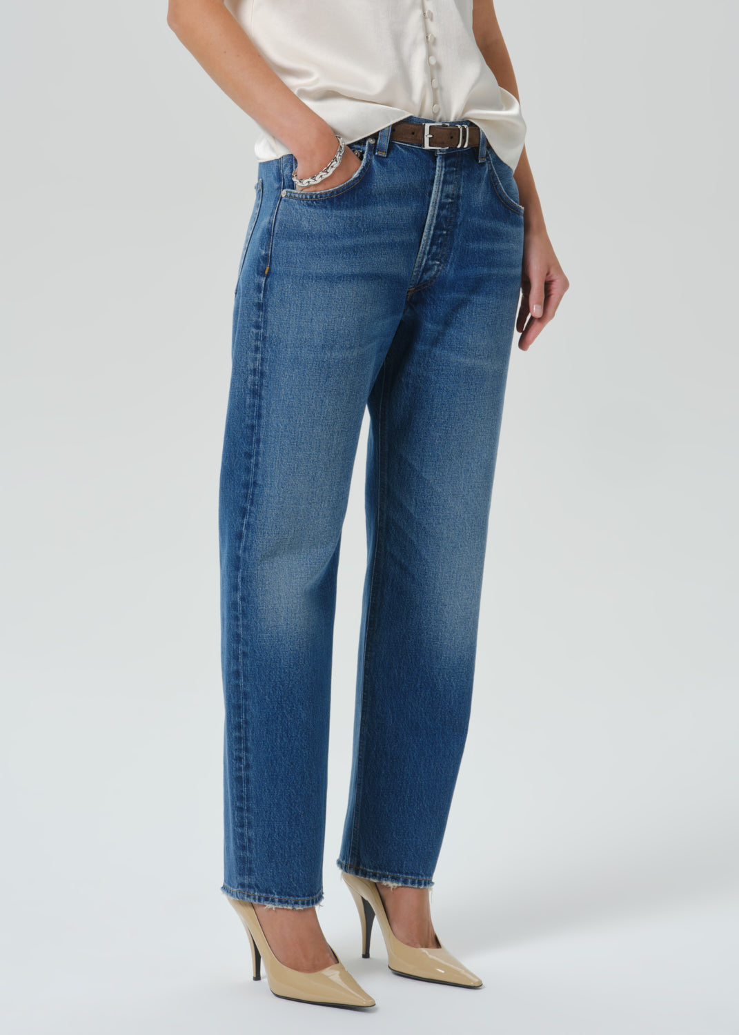 A model wearing a pair of medium-wash blue straight-leg jeans with a silver chain detail on the pocket, styled with a cream satin blouse and beige pointed-toe heels against a grey background.
