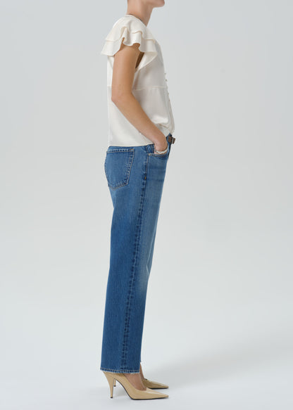 A side-profile studio shot of a model wearing blue straight-leg jeans, a cream ruffled sleeveless top, and beige high heels against a grey background.