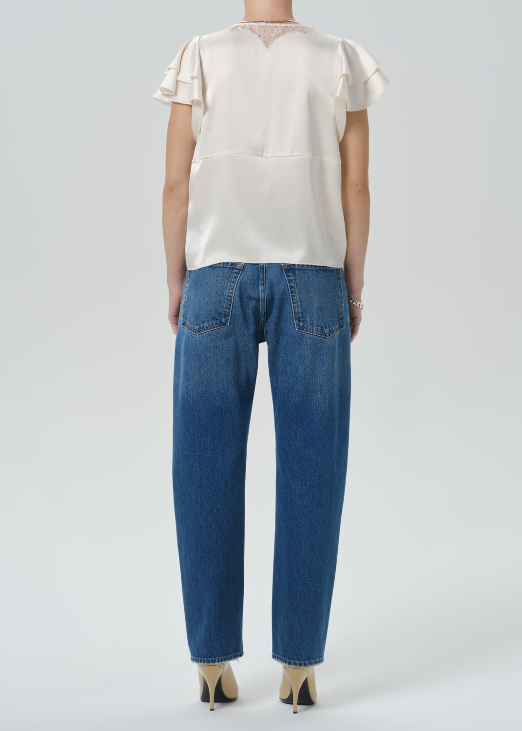 A model shown from the back wearing a cream satin short-sleeve blouse with ruffle details and blue jeans, set against a grey background.