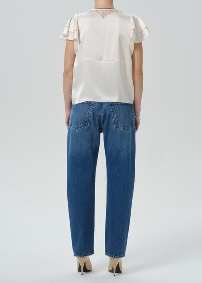 A model shown from the back wearing a cream satin short-sleeve blouse with ruffle details and blue jeans, set against a grey background.