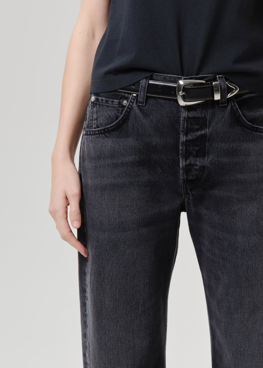 Close-up of a person wearing black denim jeans with a black belt featuring a silver buckle, paired with a black top against a grey studio background.