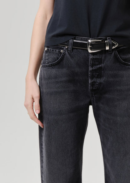 Close-up of a person wearing black denim jeans with a black belt featuring a silver buckle, paired with a black top against a grey studio background.