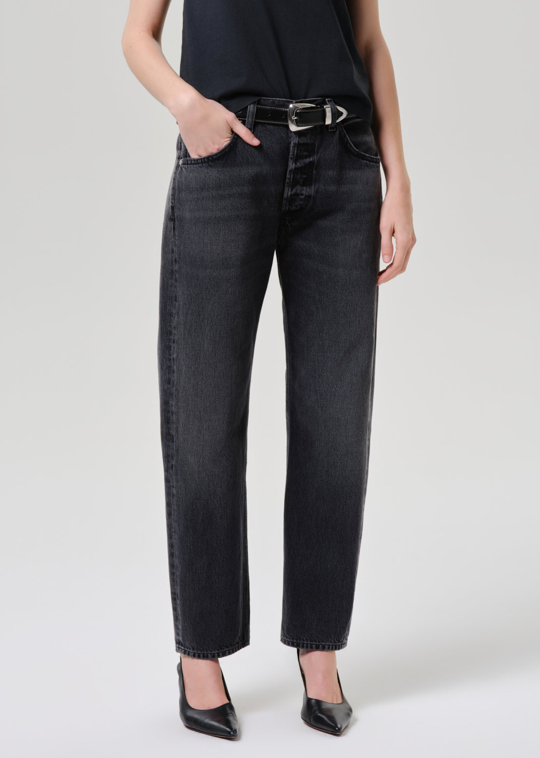 A model wearing a pair of black washed straight-leg jeans with a black belt, paired with a black top and black pointed-toe heels, against a grey  background.