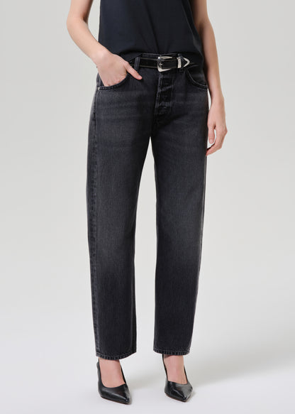 A model wearing a pair of black washed straight-leg jeans with a black belt, paired with a black top and black pointed-toe heels, against a grey  background.