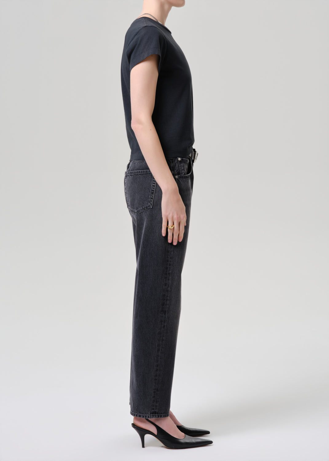A side-profile studio shot of a model wearing a black short-sleeve t-shirt, black straight-leg jeans, and black slingback heels against a grey background.