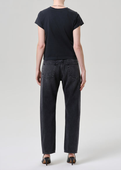 A model shown from the back wearing a black short-sleeve t-shirt, black washed straight-leg jeans, and black pointed-toe heels against a grey  background.