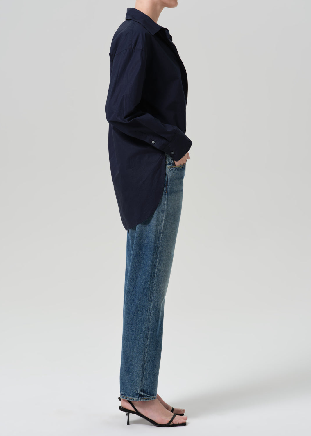 Person wearing a navy long sleeve button front blouse with medium wash straight leg jeans viewed from the side on a grey background