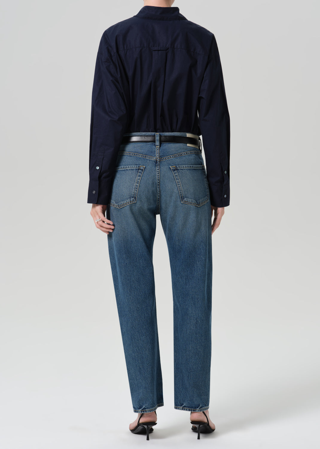 Person wearing a navy long sleeve button front blouse with medium wash straight leg jeans viewed from the back on a grey background