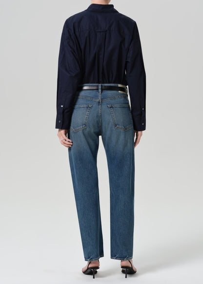 Person wearing a navy long sleeve button front blouse with medium wash straight leg jeans viewed from the back on a grey background