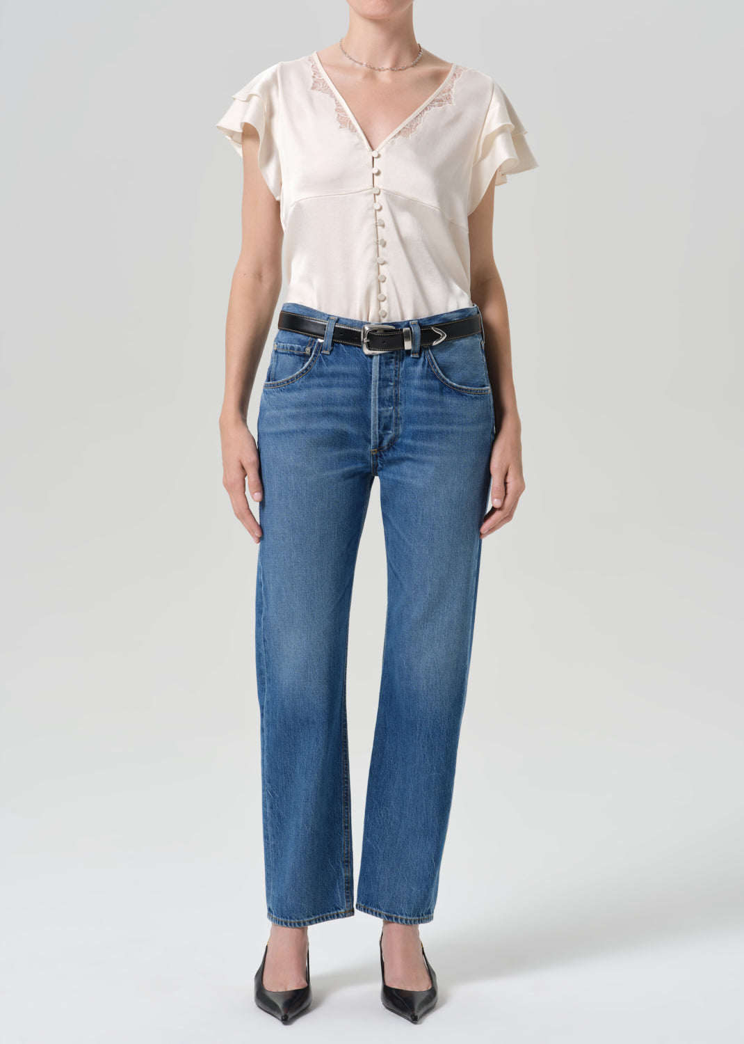 Person wearing medium wash straight leg jeans with a white short sleeve blouse on a grey background