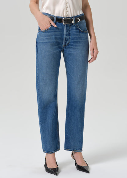 Person wearing medium wash straight leg jeans with a white short sleeve blouse and black pointed toe heels on a grey background