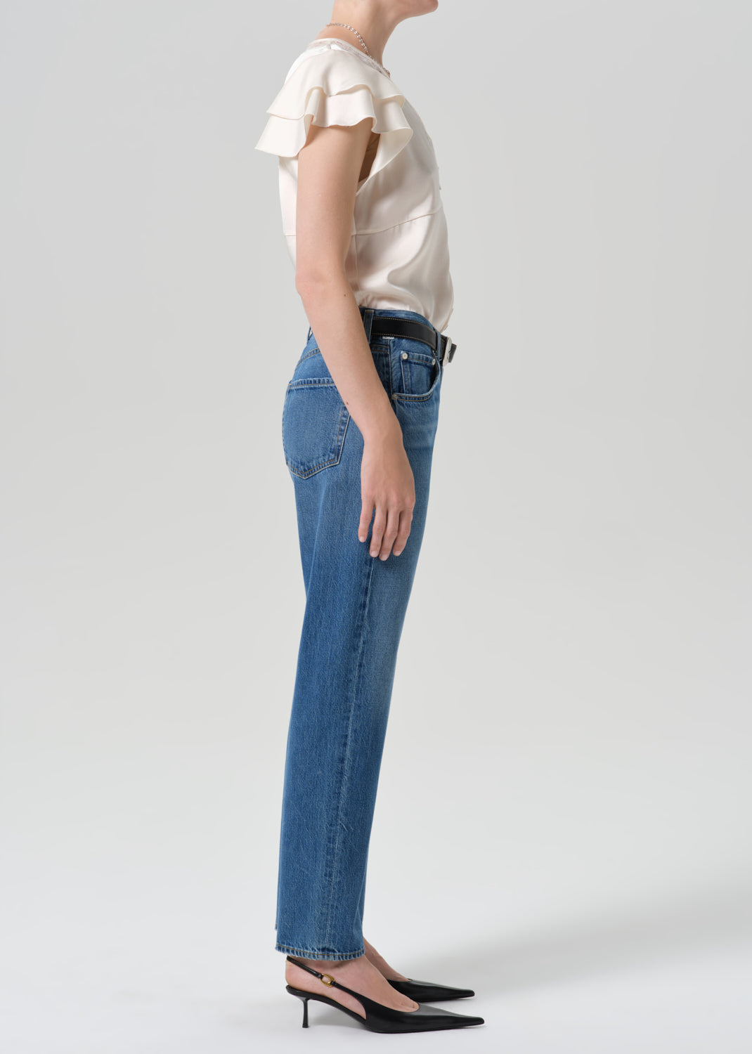 Person wearing medium wash straight leg jeans with a white short sleeve blouse viewed from the side on a grey background