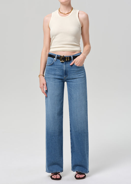 A model wearing a pair of wide-leg blue jeans with a black belt, paired with a cream ribbed tank top and black sandals, shot against a grey background.