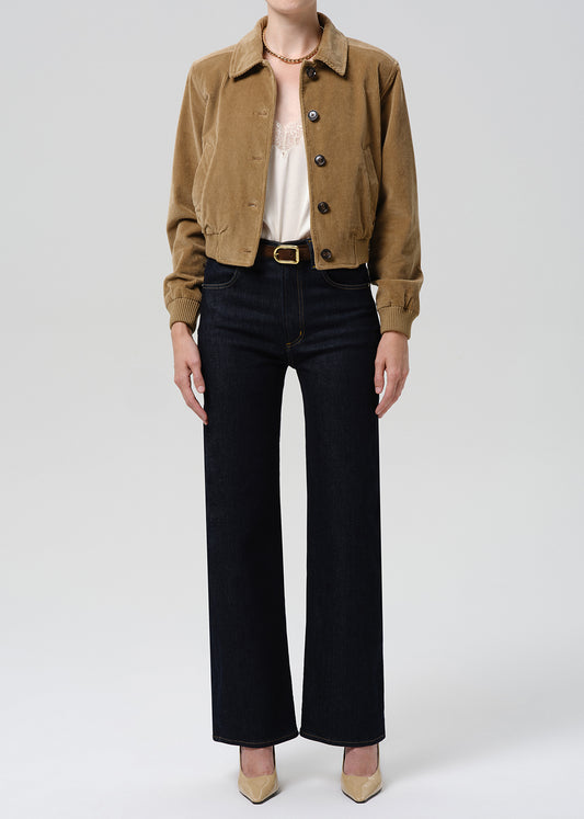 A model is shown from the neck down wearing a cropped tan corduroy jacket, a cream lace top, dark wide-leg jeans, and beige heels against a grey background.
