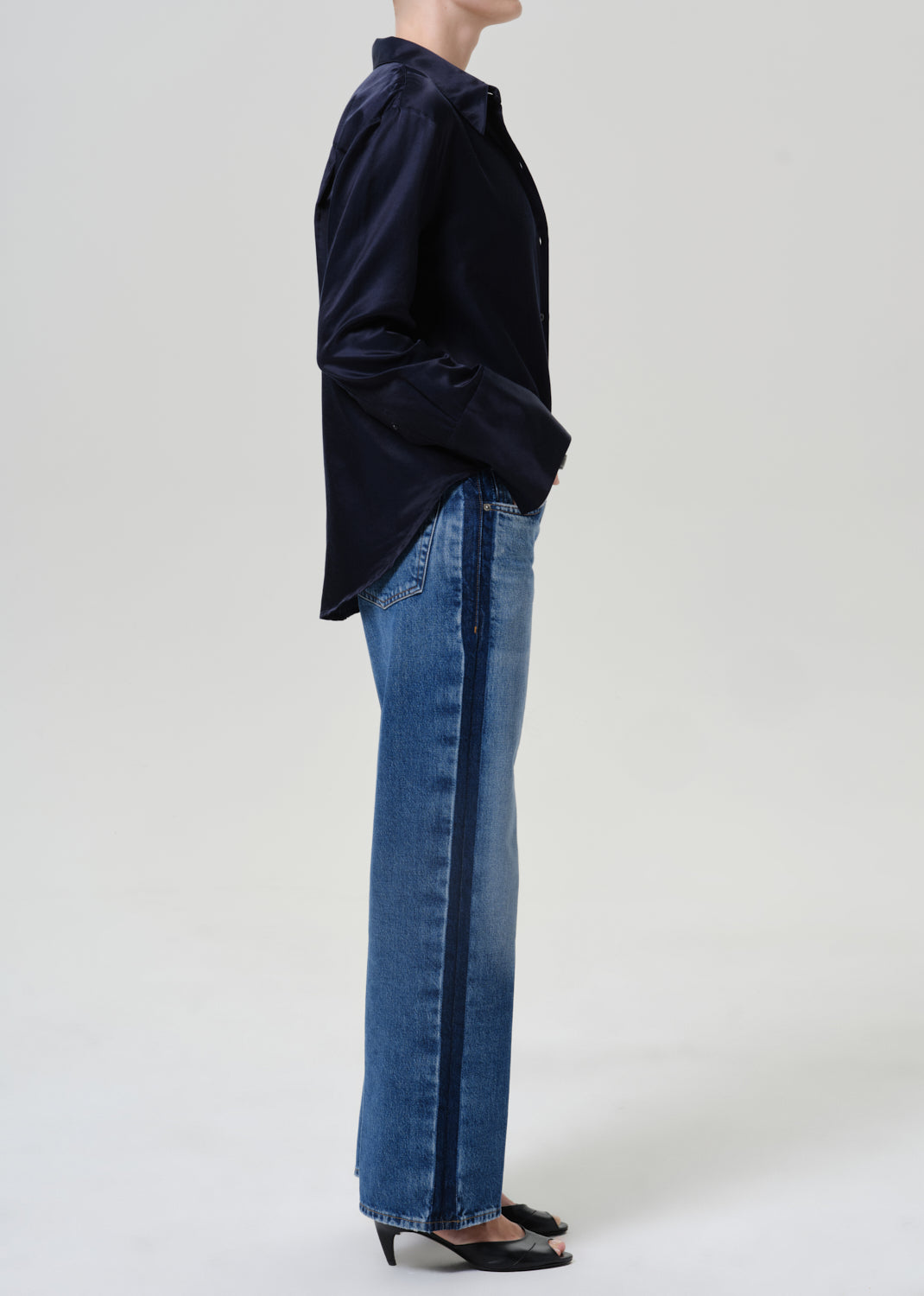 Woman wearing medium blue wide leg jeans with a dark navy button-up shirt viewed from the side on a grey background.