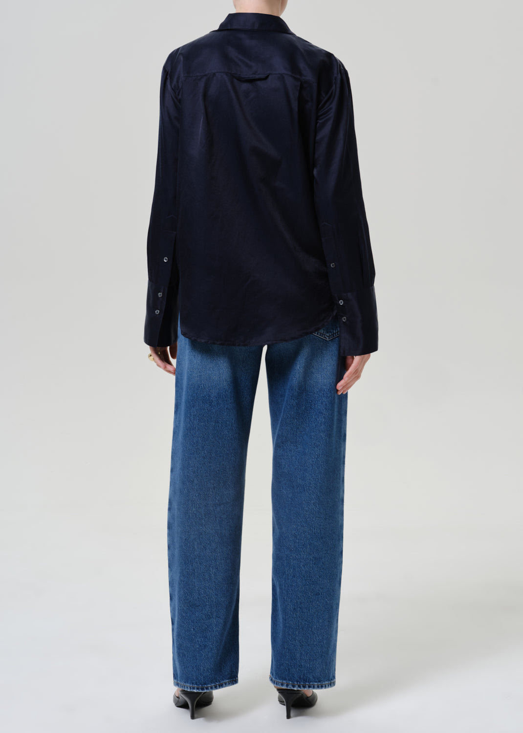 Woman wearing medium blue wide leg jeans with a dark navy button-up shirt viewed from the back on a grey background.