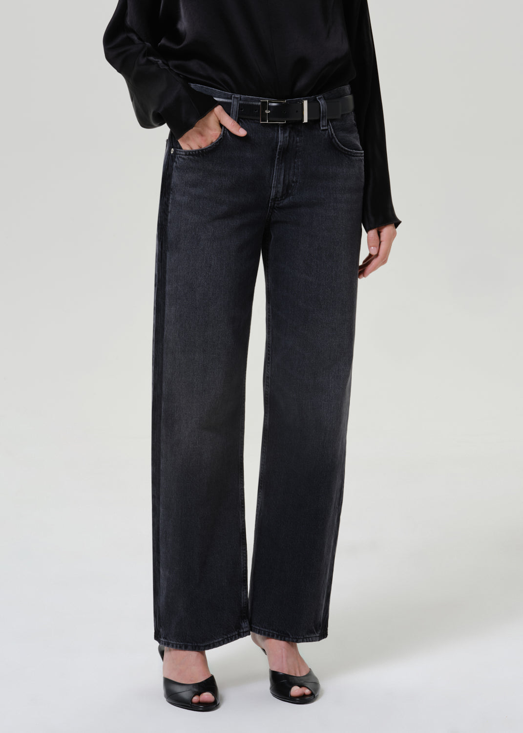 Woman wearing dark grey wide leg jeans with a black draped neck top on a grey background.
