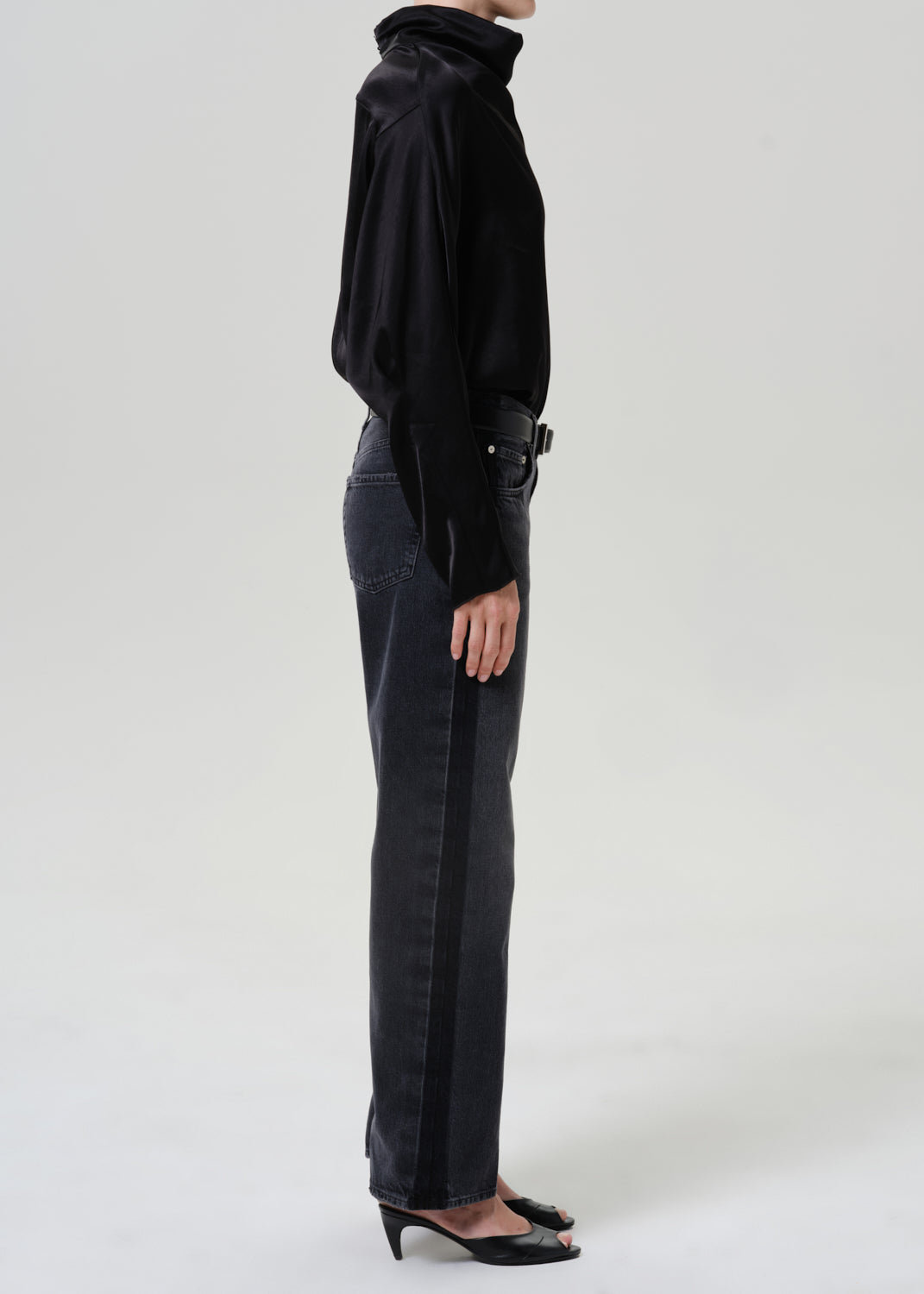 Woman wearing dark grey wide leg jeans with a black draped neck top viewed from the side on a grey background.